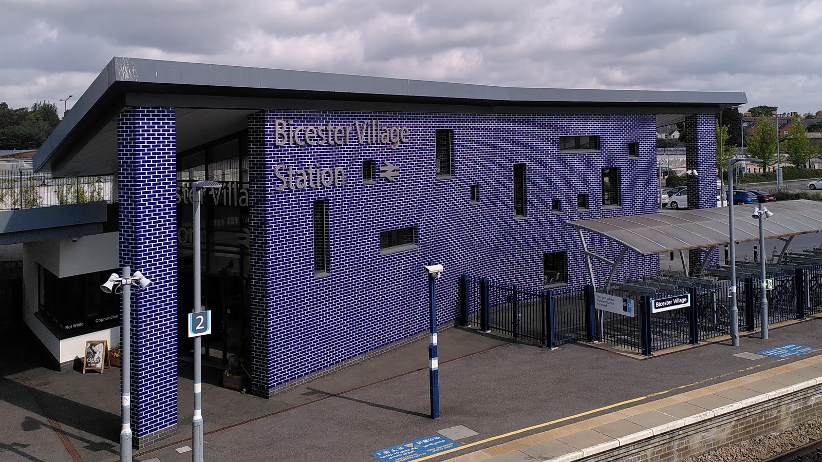 Bicester Village railway station platforms, tracks, and footbridge under a blue sky.