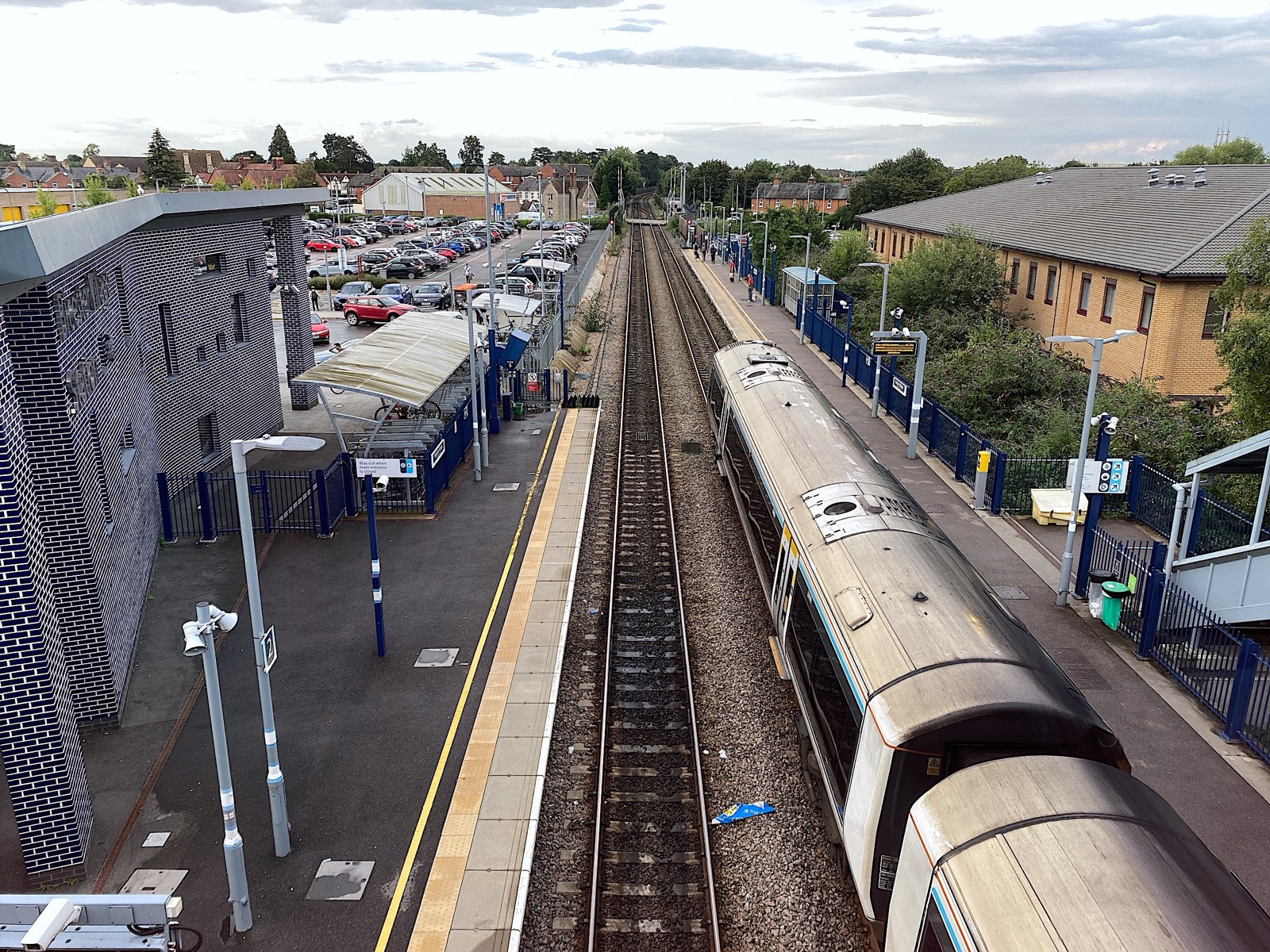 Elevated view from the footbridge at Bicester Village railway station showing platforms, tracks, modern station building, and car park, photographed in August 2023.