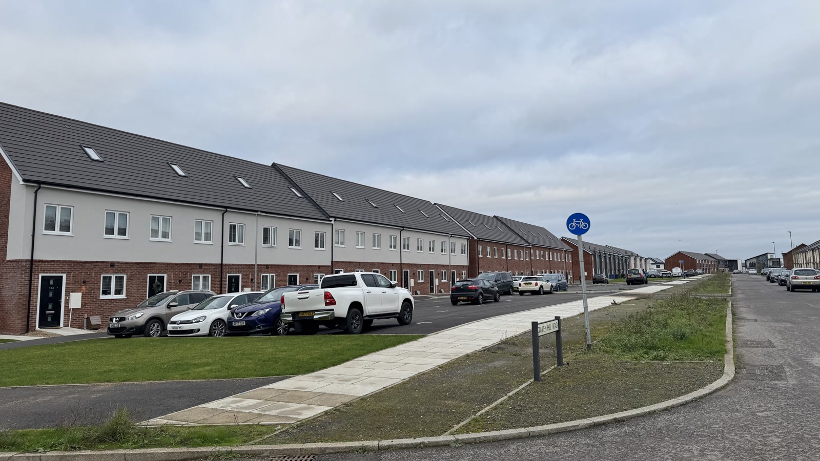 Graven Hill Road in Bicester showing newly constructed residential terraces, paved footpaths, and designated cycle routes, photographed in November 2024.